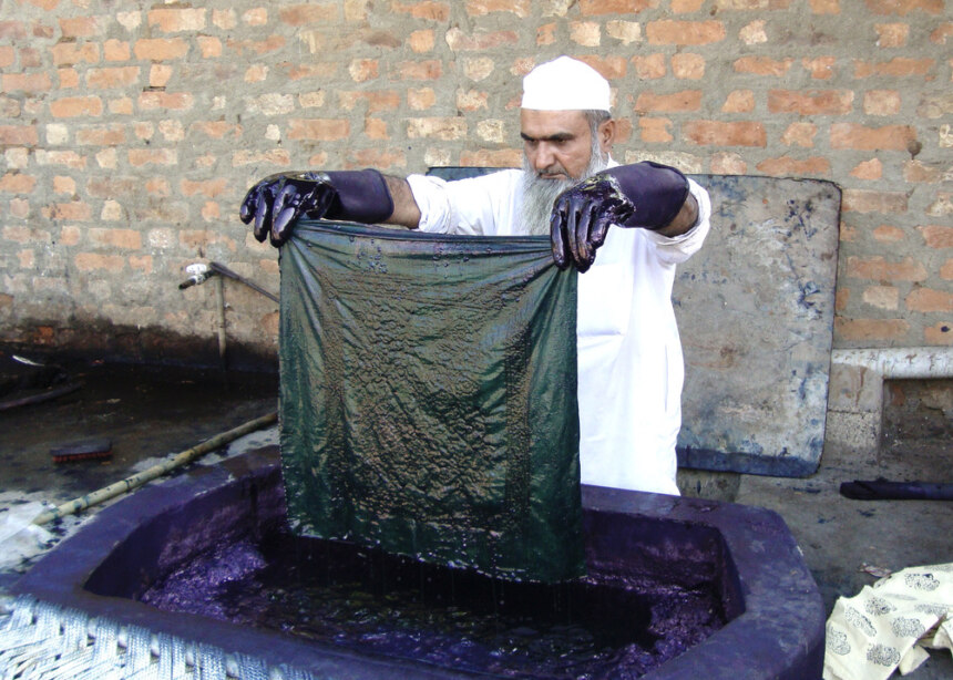 A man wearing white clothes dips a piece of fabric in indigo dye bath. 