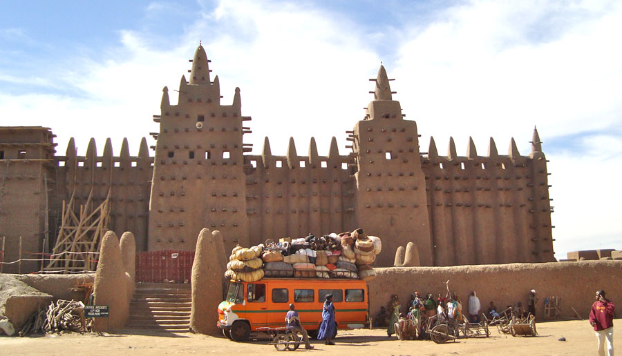 Enormous adobe block mosque with two towers and a bus in front.