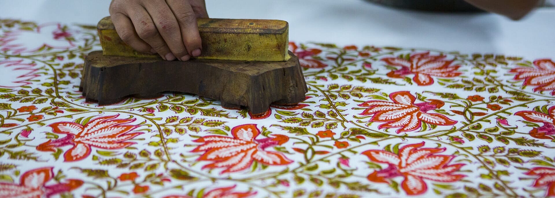 Man holds a carved wooden block and stamps red flowers onto a cotton cloth.