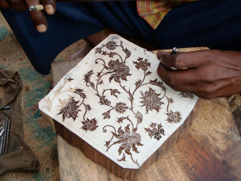 Detail of worker's hand behind the scenes, carving a wooden block with a flower pattern.