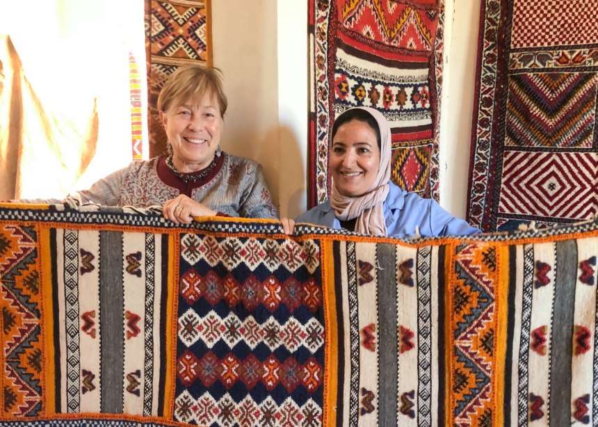 Two smiling women hold up a handwoven Morocco carpet.