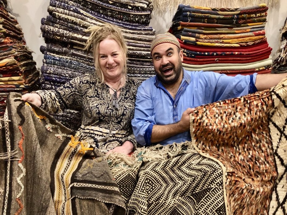 A smiling man sits and shows rugs to a blonde tourist, in front of stacks of carpets.